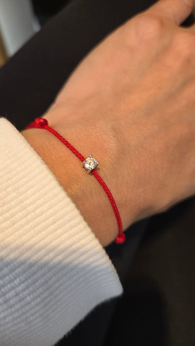 A close-up shot focuses on a woman's wrist adorned with a thin red string bracelet featuring a small, square-set diamond charm. The bracelet is worn on the left wrist, partially obscured by the sleeve of a white ribbed sweater. The background is dark and out of focus, suggesting an indoor setting. There is no identifiable information about the location in Gwangmyeong, South Korea.