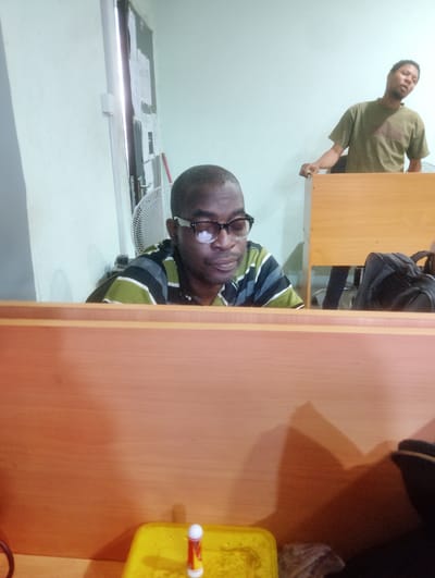 In Palmgrove, Nigeria, within what appears to be an office or study space, two individuals are captured in a photograph. The foreground is dominated by a wooden partition, obscuring the lower portion of the scene. On the left, seated behind the partition, is a Black man wearing glasses and a striped shirt. He is looking down, seemingly engaged in an activity on a surface not fully visible. To the right, standing and leaning on a wooden desk or counter, is another Black man in a green t-shirt. He is looking towards the camera and appears to be speaking or gesturing. Behind him, a black backpack is visible. The environment includes a white wall, a ceiling fan, and a notice board with papers affixed to the wall near a doorway. A yellow container with a small object, possibly a glue stick, is in the lower right corner of the frame.