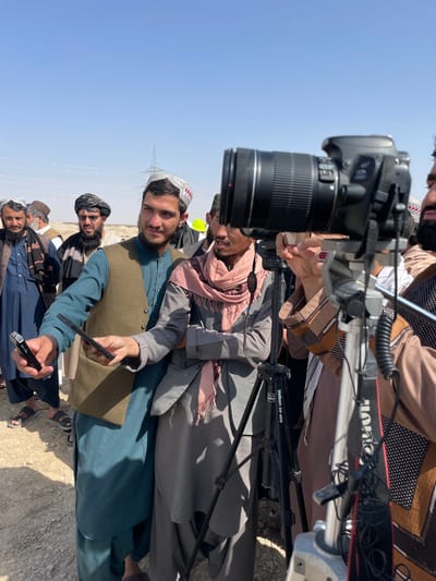 In Pir Dust, Afghanistan, a daylight outdoor scene depicts a gathering of men in an arid, open area. In the foreground right, a black Canon 700D DSLR camera with a large lens is mounted on a grey Velbon DV-400 tripod, pointing towards the center of the frame. The arm and shoulder of a man in light brown traditional attire are partially visible next to the camera.
Central to the image is a young man wearing a teal shalwar kameez, a light brown vest, and a white and red patterned cap. He holds a black smartphone horizontally in his left hand and a black rectangular device, likely a portable microphone or recorder, in his right hand, extending both forward. He looks towards his right. To his immediate right stands another young man in a grey shalwar kameez, a grey textured vest, and a pink scarf wrapped around his neck, with his arms crossed. He also looks in the same general direction.
Behind these central figures, a group of approximately ten to fifteen men are observed. They wear traditional Afghan clothing, including various colors of shalwar kameez, turbans, and caps; many have beards. One prominent figure in the mid-ground wears a black turban, dark traditional clothing, and a significant beard.
The ground is dry, light brown dirt or sand. The sky is clear and light blue, suggesting bright daylight. In the distant background, a large metal lattice power transmission tower is visible under the clear sky.