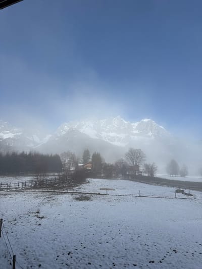 An outdoor panoramic image depicts a winter landscape in Going am Wilden Kaiser, Austria. The upper third of the frame presents a clear, bright blue sky. Directly beneath, a dense stratum of white mist or low clouds horizontally obscures the middle ground, partially concealing the snow-covered, rugged peaks of the Wilden Kaiser mountain range. Below the mist, a rural scene unfolds. Several bare deciduous trees and dark coniferous trees are interspersed among snow-dusted fields. A cluster of traditional wooden buildings, consistent with chalets, is visible in the mid-distance near the base of the mountains. The foreground consists of expansive fields covered in a thin layer of fresh snow, with patches of darker ground visible. Multiple dark wooden fences delineate these fields, and a wooden feeding trough or signpost stands in a field on the right. No individuals, animals, or dynamic actions are depicted. A dark architectural element partially frames the top-left corner, indicating the image was captured from an interior vantage point looking outward.