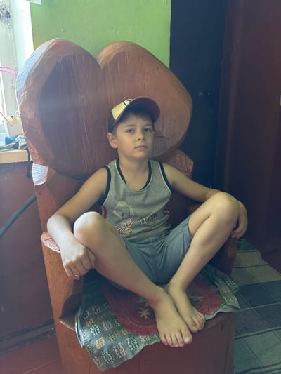 A young boy, wearing a gray tank top with black and white trim and blue shorts, is lying down on a wooden bench. He is wearing a baseball cap backwards. The boy is looking at the camera with a neutral expression. His legs are bent and crossed, and his arms are resting on his legs. A patterned cushion or blanket is under his legs.
The bench appears to be part of a wooden structure with large, rounded, heart-shaped elements. The background shows a green wall on the left and a wooden wall with a tiled floor on the right. The lighting suggests it is daytime, likely indoors. The setting is rustic, possibly a porch or a room with wooden furniture. There is some visible text on the boy's shirt: "1998.900", and possibly some other partial text. The overall scene evokes a casual, relaxed moment.