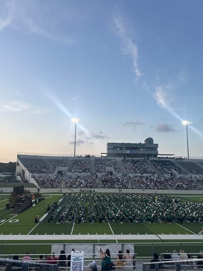 The image captures a graduation ceremony at Dragon Stadium in Southlake, Texas, United States of America. Graduates wearing green gowns and white caps are seated in rows on the green football field. The stands are filled with spectators. In the upper left, the sky is blue with some clouds and bright stadium lights. The structure bearing the name "Dragon Stadium" is visible, along with a Methodist logo. A fence and some people are in the extreme right foreground. A QR code is affixed to the fence.