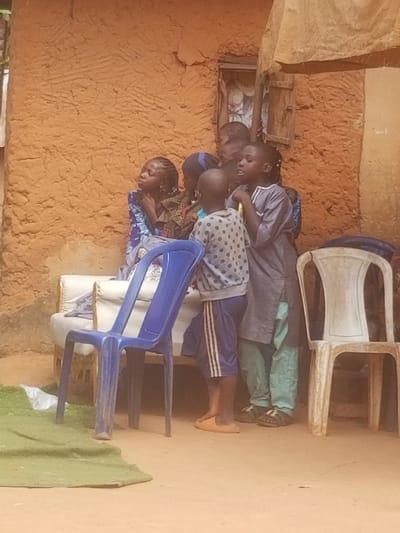 This image depicts a gathering of approximately 6-7 children outdoors in Ochadam, Nigeria, positioned against an unpainted, textured orange-brown mud wall. A small, open rectangular window with a dark wooden shutter is visible on the wall in the upper right. An off-white fabric or tarp is draped above and partially hangs into the upper right of the frame. The ground is light brown dirt.
In the center-left, a female child with braided hair, wearing a patterned blue and purple garment, sits on a white upholstered bench, gazing upwards and to the left. Adjacent to her and standing are several other children. A male child in the foreground, wearing a grey polka-dotted long-sleeved shirt and blue shorts with white stripes, stands in orange slides, facing right. To his right, another male child in a grey long-sleeved tunic over light green trousers and black sandals appears to be speaking or singing, with his mouth open, looking towards the right of the frame. Other children are partially visible behind these two, observing in the same general direction.
In the immediate foreground, a blue plastic monobloc chair stands, facing right, partially obscuring the lower bodies of the standing children. To the right, a white plastic monobloc chair stands, facing left. A folded or bunched green fabric lies on the ground in the lower-left corner.