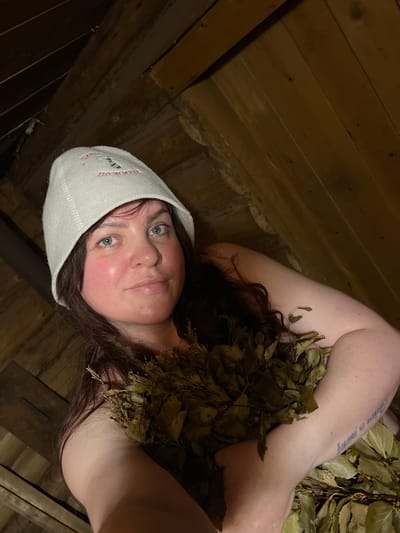 A woman lies among the leaves in what appears to be a sauna. She is fair-skinned with brown hair and blue eyes, wearing a white sauna hat with red lettering. There is a tattoo that says "Believe in yourself" on her arm. The background shows wooden planks, typical of sauna interiors. The image was taken in a sauna, and the presence of the sauna hat and leaves suggests the woman is engaging in a traditional sauna ritual. There is no indication this image was taken in the city of Protasy, Russia.