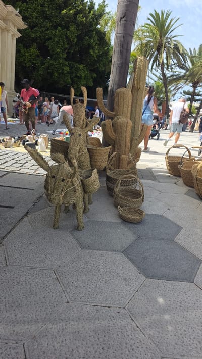 In Almuñécar, Spain, on a bright, sunny day, a street market is in full swing. The ground is paved with large, hexagonal grey stones. In the foreground, two decorative straw donkey figures stand with baskets on their backs, resembling pack animals, next to a woven straw cactus sculpture. Surrounding these items are various woven straw baskets of different sizes, some displayed on the ground, and others stacked. In the background, a bustling crowd of people walks and browses the market stalls. Palm trees and lush green trees frame the scene under a clear blue sky. A man in a red shirt and dark cap, holding a small dog, stands near the left side of the image, engaged in conversation. To the right, a woman in denim shorts and a white top walks away from the camera. Other shoppers are visible in the distance, creating a lively atmosphere.