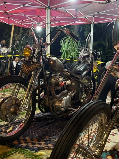 The image shows a collection of vintage motorcycles on display under a red and white tent, potentially at a motorcycle exhibition or gathering in Banjar Anyar, Indonesia. The motorcycles are the primary subjects, showcasing their intricate designs and chrome details. People are standing near the motorcycles, appearing to be observing or discussing them. The environment includes vegetation and what appears to be a patterned textile or rug placed near one of the motorcycles.