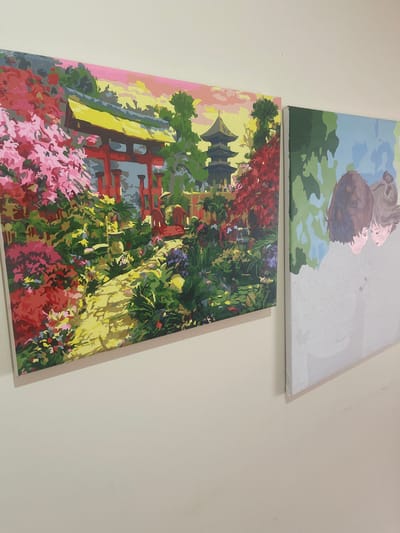This image shows two unfinished paint-by-number canvases hanging on a light-colored wall. The canvas on the left depicts a vibrant scene with a red torii gate, a pagoda, and lush pink and red foliage. A stone path winds through the garden. The canvas on the right shows a close-up of two figures, possibly a couple, with their heads close together. They are rendered in muted tones, with numbers still visible on the canvas, indicating it is incomplete. The scene is indoors, as suggested by the lighting and the way the canvases are mounted on the wall. There is no indication that this scene is located in São João das Lampas, Portugal.