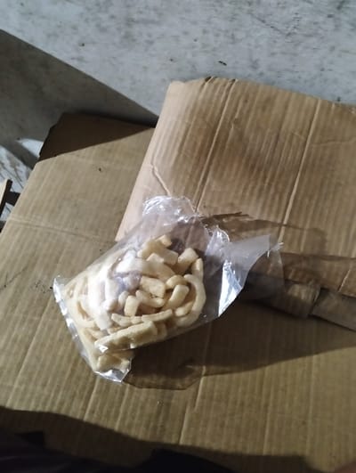 The image shows a bag of light-colored snacks, possibly chicharron or similar fried pork rinds, placed on top of corrugated cardboard. The snacks are cut into irregular, bite-sized pieces and are enclosed in a transparent plastic bag. The bag appears to be partially open, revealing the contents.
The scene is set in what seems to be an indoor, dimly lit environment. The background consists of a concrete wall with a noticeable shadow cast from an unseen object on the left side of the frame. The cardboard surfaces suggest a storage or shipping area. There is some faint handwriting visible on the lower left cardboard, with numbers like "130", "12", and "24" discernable.
The lighting suggests it could be nighttime or a poorly lit indoor space. The overall impression is that of a casual setting, possibly a home or a small business storage area in Palohgadeng, Indonesia. No people are visible in the image, and the focus is solely on the bag of snacks and the surrounding cardboard. The mood is neutral, with no strong emotional cues conveyed.