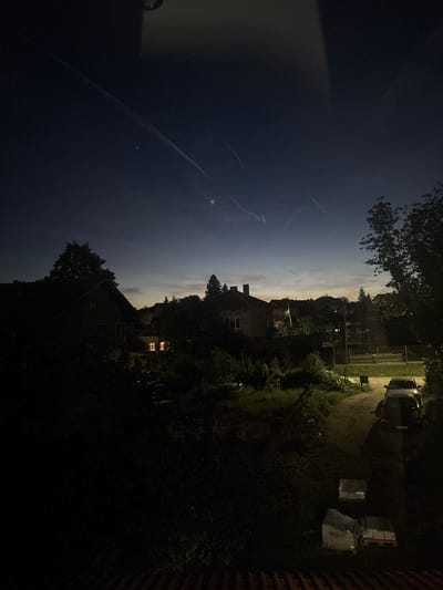 The image shows a nighttime scene of a residential area. The sky transitions from a dark blue on the left to a lighter, cloudy sky above the horizon. A contrail is visible in the dark blue sky. The foreground contains silhouetted trees and buildings. In the middle ground, there is a house with illuminated windows. Further to the right, a car is parked near stacks of covered building materials. The exact location in Petarch, Bulgaria cannot be identified from the image alone.