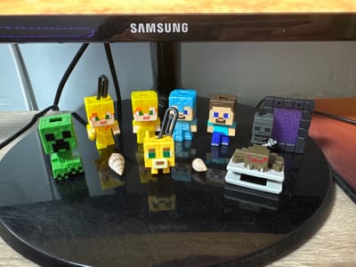 The image shows a collection of Minecraft-themed figurines arranged on a black circular surface, which appears to be the base of a monitor. The figurines include a green Creeper, two yellow characters, a blue character, a character with brown hair, a skeletal figure, a purple Nether Portal, and a spider. Two small seashells are also present, positioned near the yellow figurines. In the background, a Samsung monitor is visible, displaying subtle LED lights. The surface on which the figurines are placed has visible scratches and dust. The scene is lit from above, creating soft shadows. The environment appears to be indoors, likely on a desk. There is no indication of the city of Pavolche, Bulgaria in the image.