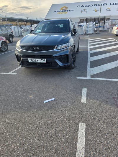 This media file shows a dark grey Geely car parked in a lot with a large building in the background. The building has a prominent sign with Russian text that reads "САДОВЫЙ ЦЕНТР" and "ЛЕМАНА ПРО". The car has a license plate that reads "T853XP 154". White painted lines on the asphalt indicate parking spaces. There are stacks of bagged goods behind the car, possibly related to the garden center. The sky suggests it is either early morning or late afternoon, with soft lighting. The scene appears to be a commercial area, likely a garden supply store. There are no people visible in the image.