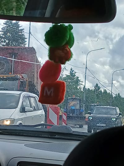 A fuzzy, red, square object with a white "M" is hanging from the rearview mirror of a car, in front of a cloudy sky. Below the object, a red and white construction barrier is visible, followed by a white minivan with its headlights on. Further in the background, a dark-colored SUV with its headlights on, a tractor, and a red truck are seen in what appears to be a traffic jam. The scene is set in Kuznetsovo, Russia.