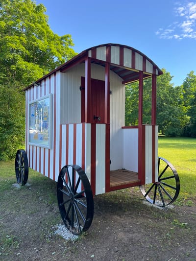 A small, red and white, wagon-like structure sits on a grassy and dirt area. The structure has a rounded, arched roof and is supported by red beams. It features two large black spoked wheels on each side, attached to the structure by chains. A window on the side of the wagon displays what appears to be a poster or information board with multiple images and text, though the text is illegible. The wagon is painted with alternating red and white horizontal stripes on its lower half. The setting is outdoors, with lush green trees and a clear blue sky visible in the background, suggesting a sunny day. The ground is a mix of grass and exposed dirt, with some scattered white gravel near the wheels. The overall scene appears to be a park or a historical site. No people are visible in the image. The time of day appears to be daytime. The location context is Narva-Jõesuu, Estonia.