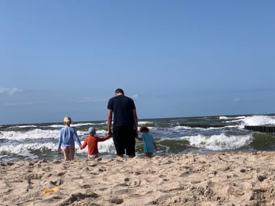 In the city of Rewal, Poland, a man and three children are standing on a sandy beach facing the Baltic Sea under a clear blue sky. The man, wearing a dark blue t-shirt and black shorts, holds the hands of two children. To his left, a young girl in a blue long-sleeved shirt and pink bottoms stands holding the man's hand and appears to be looking out at the sea. Next to her, a young boy in an orange t-shirt and a light blue cap holds the man's hand. To the man's right, another young boy in a light blue t-shirt stands holding the man's other hand. The children are wearing swim trunks or bottoms. The waves of the sea are breaking on the shore, and wooden breakwaters are visible in the water. A yellow plastic shovel lies in the sand in the foreground.