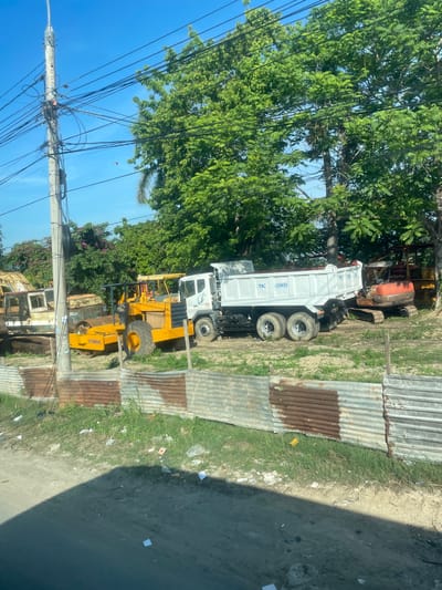 A construction site in Diên Lạc, Vietnam, is visible with several heavy machinery vehicles. In the foreground, a yellow Dynapac roller is parked, and behind it, a white dump truck with blue lettering that reads "79C 08681" is present. Further to the left, a white and rust-colored excavator is partially obscured by a utility pole and overhead wires. To the right of the dump truck, an orange excavator is also parked. The site is bordered by a corrugated metal fence, some sections of which are rusted. Trees and foliage are abundant in the background, and the sky is clear blue. The ground is a mix of dirt and grass, with scattered debris.