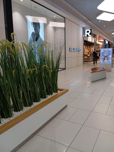 The image depicts an indoor scene within a shopping mall. A person is walking in the background, and various shop signs are visible, including a prominent "R" logo and the word "RESERVED" in illuminated letters. Artificial greenery with yellow accents is placed in the foreground, contained within a planter. A large digital display showing a person's portrait is also visible. The setting appears to be a modern, well-lit shopping environment. The time of day is not discernible, and there are no indications of weather. The scene suggests a typical day at a retail establishment in Wołomin, Poland.