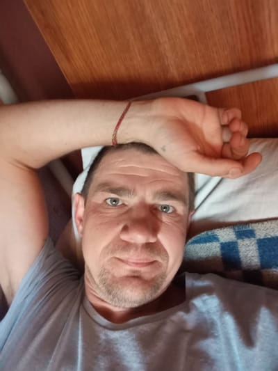 A close-up, eye-level, selfie-style shot captures a fair-skinned Caucasian man with short, dark hair and stubble on his face. He is lying down on a white pillow and a blue and white checkered blanket, his head turned towards the camera. His right arm is raised, with his hand resting on his forehead, revealing a red and green string bracelet on his wrist. He is wearing a light gray t-shirt. The background consists of a wooden surface, possibly a shelf or headboard, with a white bar running horizontally. The lighting appears to be natural, coming from the front and slightly above the subject. The man is looking directly into the camera with a neutral expression. The setting is suggestive of an indoor environment, possibly a bedroom or a resting area. The image does not contain any specific landmarks or elements that would indicate its precise location within Solonytsivka, Ukraine.