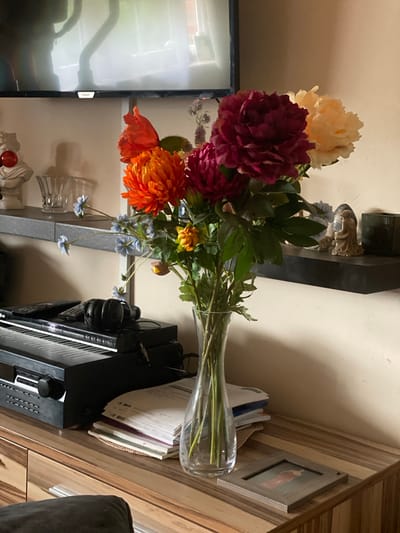 The image shows a collection of artificial flowers in a clear glass vase. The flowers are a mix of colors, including deep red, orange, and cream, with green foliage. Behind the flowers, on a shelf, are several decorative figurines and a dark, angular object. To the right of the flowers, a wooden surface displays a stack of papers and a small framed picture. Further to the right, a black electronic device with buttons and a dial is visible, along with a pair of black headphones. In the background, a beige wall is present, and to the left, a portion of a television screen is visible. The lighting suggests it is daytime, and there are no indications of weather conditions or specific emotions. No visible text is present in the image, other than a small "Philips" logo on the television.