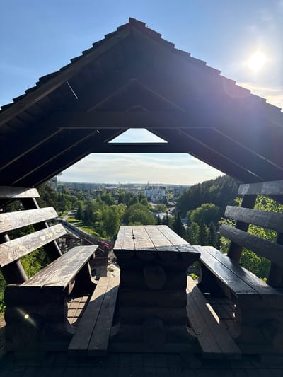 The image depicts a view from within a wooden structure, possibly a lookout tower, in Belokurikha, Russia. The composition is framed by the dark, angular timbers of the structure itself, with the bright sun visible at the top left. A rectangular opening in the wooden construction provides a vista of the surrounding landscape.

In the distance, a cityscape is visible, characterized by buildings of varying heights. The foreground and middle ground are dominated by lush green trees, suggesting a park or forest area within the city. A pedestrian bridge under construction is visible on the right side of the view.

The wooden structure appears weathered and rustic, with sunlight casting sharp shadows across its surfaces. The overall impression is one of being elevated and looking out over a combination of urban and natural landscapes.