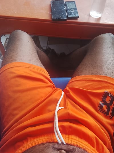 The image shows a person's legs and lap from a low-angle perspective, wearing bright orange shorts with a white drawstring. The shorts have a black graphic logo on the right leg. The person is seated at an orange plastic table, where two cell phones and a partially filled glass of water are visible. The lighting suggests it is daytime, and the overall scene appears casual, possibly outdoors or in a brightly lit indoor space. The location is identified as Orodo, Nigeria. The cell phones appear to be older models, one possibly a Tecno phone. The texture of the person's legs indicates a warm climate, given the visible hair and the casual attire.
