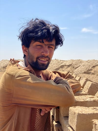 A man stands in Deh-E Bagh, Afghanistan, leaning against a stack of sun-dried mud bricks. He has dark, curly hair, a short beard, and is wearing a tan collared shirt over a patterned undershirt. The background shows more stacks of similar bricks under a clear blue sky.