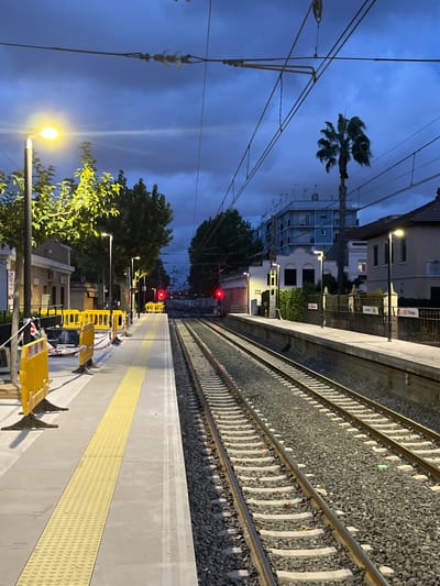 The image depicts an unoccupied scene at the Picanya train station in Picanya, Spain, observed during twilight or night under a dark, cloudy sky. Two sets of railway tracks, laid on gravel ballast, run through the center of the frame, receding into the distance where two illuminated red signal lights are visible. Overhead electrical catenary lines crisscross against the sky.

On the left side, a concrete train platform is present, featuring a prominent yellow tactile paving strip. Several bright yellow temporary barriers, some connected by red and white striped tape, are positioned on this platform, suggesting a demarcated area. A street lamp in the immediate foreground illuminates the platform and the foliage of nearby trees. A low-rise building is partially visible behind the trees on the left.

On the right side, a second concrete platform is visible, also illuminated by a street lamp. A black sign with white text displaying "D. Picanya" is affixed to a fence adjacent to this platform. Beyond the right platform, in the background, a tall palm tree stands in front of a multi-story light blue building equipped with numerous rooftop antennas. Additional residential-style buildings with tiled roofs are also visible. No individuals or active interactions are observed in the scene.
