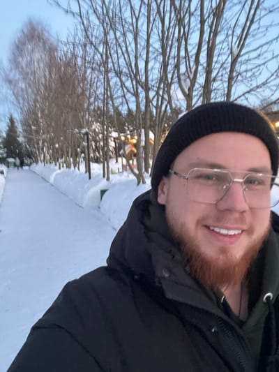 A selfie of a smiling man with a red beard wearing glasses and a black beanie, taken outdoors in a snowy environment. Trees with bare branches line the background, with a dusting of snow visible on the ground. He wears a dark, puffy jacket. The photo is taken at a slight angle, with the man's face positioned towards the upper right corner of the frame. Soft natural light illuminates the scene.