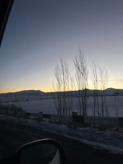 A winter landscape is viewed from a car window, with bare trees lining a snow-covered roadside. Beyond the trees, a distant, snowy mountain range meets a pale, hazy sky transitioning from light to dark. A guardrail is visible between the car and the landscape. The car's side mirror is partially visible in the lower right corner. The sun appears to be setting or rising, casting a soft, golden glow on the horizon.