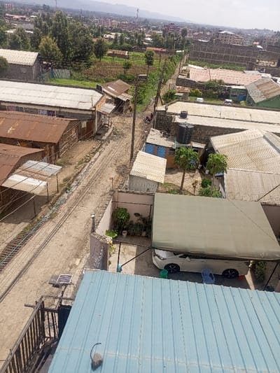 An elevated view captures a semi-urban landscape in Kiamaina ward, Kenya, under daylight conditions. The scene is dominated by a central unpaved dirt road, flanked by various residential and commercial structures. To the immediate left of the road, a parallel, narrower track or drainage channel runs along multiple buildings featuring corrugated metal roofs, some exhibiting rust. One larger structure with a white corrugated roof and visible windows on an upper floor stands prominent on the left.

On the right side of the dirt road, a concrete wall topped with barbed wire encloses a property. Within this enclosure, a white multi-purpose vehicle is parked beneath a large, light-colored fabric canopy supported by dark metal poles. Two small plastic chairs, one green and one blue, are situated near the vehicle. Adjacent to the carport, a small courtyard area contains potted plants. Further along this side of the road, a cluster of buildings with diverse roofing materials, including corrugated metal and flat concrete, are visible. One building features a large black water storage tank on its roof.

Utility poles with multiple power lines extend along the dirt road. A single individual on a red motorcycle is observed on the dirt road in the mid-distance, traveling away from the viewer.

In the foreground, the edge of a light blue corrugated metal roof is visible, with a small dark object resting upon it. To the left foreground, a dark metal railing structure and a solar panel unit mounted on a pole are present. The background reveals a progression of additional buildings, interspersed green vegetation, and distant hills or mountains under a hazy sky, indicating further urban and natural terrain.