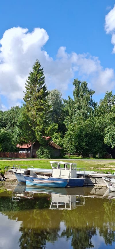 This is a vertical, eye-level, outdoor photograph of a waterfront scene in Valma, Estonia. The foreground features the tranquil, reflective surface of a body of water, likely a lake or river, with the mirrored image of the sky and surrounding trees. Moored to a concrete dock in the middle ground are two boats. The larger boat, to the right, is white with a blue hull and features a cabin with windows. Beside it, to the left, is a smaller, bright blue boat with a white interior and a railing. The background is dominated by lush, green trees and foliage, with a few wooden structures, possibly cabins or sheds, partially visible amongst the trees. A wooden fence runs along the edge of the property. In the distance, a tall evergreen tree stands out against a bright blue sky dotted with white, fluffy clouds. The overall impression is one of peaceful rural life by the water.