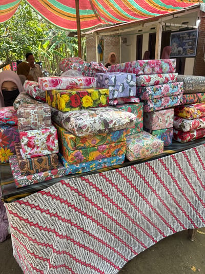 In Kruengseupeng, Indonesia, a street vendor displays stacks of wrapped gifts under a colorful striped awning. The gifts are wrapped in various patterned papers, featuring floral designs, penguins, and cityscapes. In the foreground, a table is draped with a red and white batik cloth. Behind the table, two individuals are visible, one wearing a black face mask and a pink hijab, and another wearing a brown hijab. A man with a light-colored shirt and a woman with a brown hijab are partially visible in the background. A framed sign, possibly indicating a wedding celebration, hangs on the wall of a building behind the vendor's stall. The environment appears to be an outdoor market or street setting, with trees and buildings visible.