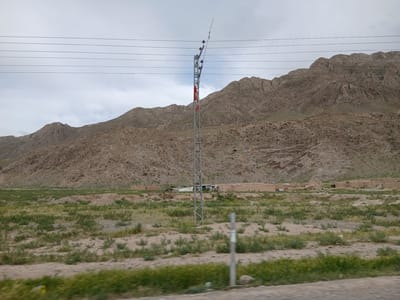 The image shows a landscape in Bostan, Pakistan, featuring mountains, a rural settlement, and utility infrastructure. The mountains in the background are arid and rocky. In the midground, there is a cluster of structures that appear to be a small village or settlement, built of adobe or similar material. A metal utility pole stands prominently in the foreground with power lines extending across the top of the frame. The foreground is a sparse, grassy field interspersed with sandy patches. A section of road is visible at the bottom of the frame.