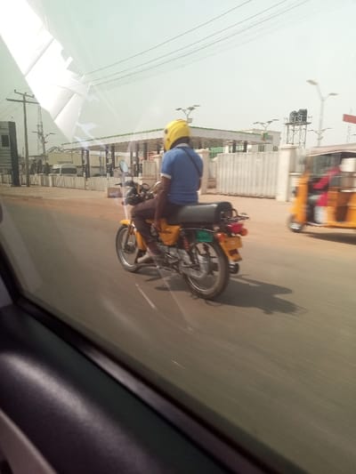 A daytime photograph, taken from inside a moving vehicle, captures a person riding a yellow motorcycle on a road in what appears to be an urban or semi-urban area. The motorcyclist, seen from behind, wears a bright yellow helmet, a blue short-sleeved polo shirt, and dark trousers. A strap, possibly from a bag, crosses his back. The yellow motorcycle has a black seat and a rear rack.
In the background, a gas station canopy is visible with what seems to be service pumps underneath. Beyond the station, white walls or fences line the roadside, and several utility poles with power lines stretch across the frame against a bright, hazy sky. To the far right, a blurry yellow motorized rickshaw, commonly known as a Keke Napep, is also in motion, suggesting traffic on the road.
The bright sunlight indicates a clear day, casting shadows of the motorcycle and rider onto the pavement. The foreground shows parts of a car's interior, including the dark dashboard and window frame, from which the picture was captured. Reflections on the car window are evident, particularly in the upper left, slightly obscuring parts of the distant background. No legible text is visible on any signs or vehicles. The scene depicts everyday transport in an active setting, consistent with the provided context of Asokoro, Nigeria.