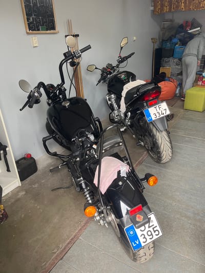 The image shows two motorcycles parked in a garage, presumably in Grīnvalti, Latvia, based on the license plates "LV 395 ZC" and "LV TF 3347" visible on the motorcycles. The motorcycles are black and appear to be similar models. The garage has a gray concrete floor and light gray walls. There is clutter in the background, including boxes, bags, and other items. Also visible is an oil container and a chalkboard on the wall. A person is lying on a bed in the upper left.