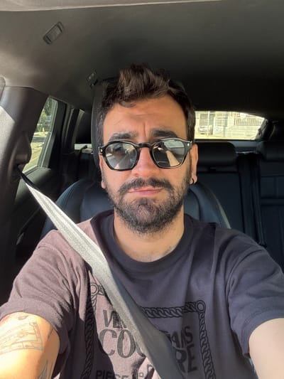 A man is seated in a car, wearing sunglasses and a t-shirt, with a seatbelt fastened. The interior of the car is black. Buildings are visible through the car windows. The location is in Asomatos, Cyprus.