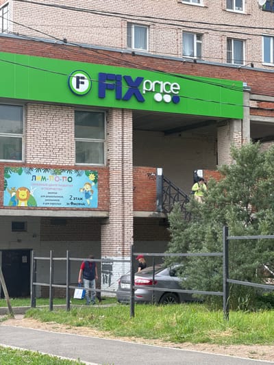 In Kuzmolovskiy, Russia, a building facade displays a bright green sign for "FIX Price" with a circular logo featuring a stylized "F". To the right of the sign, a smaller banner advertises a "Family Center for Development and Creativity" named "LIM-PO-PO", with illustrations of children and animals. The banner also indicates that classes are held on the 2nd floor behind Fix Price.
Several people are visible around the building. A person in a bright yellow jacket appears to be working on the upper part of the building. Near a parked silver car, a woman in a red hat is standing. Further down, a man in a blue vest and red shirt is holding a white bag, possibly near an entrance or walkway. The scene suggests a typical daytime activity in an urban or suburban setting, with clear weather. Power lines run vertically along the brick building. The overall impression is of a commercial area with community services.