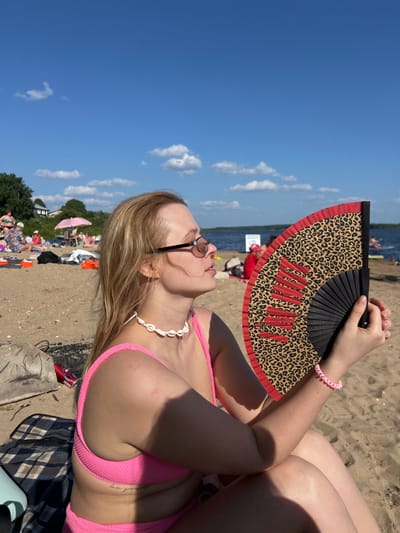 A young woman with blonde hair and sunglasses sits on a sandy beach, holding a black and tan leopard print fan with red accents. She is wearing a pink bikini and a shell necklace. The background shows a body of water, a few people on the beach, and a blue sky with scattered clouds. The location is the city of Ust'ye, Russia.
