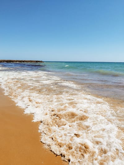 An eye-level, medium shot captures a bright, sunny day at a beach in Rota, Spain. The sky is a clear, vibrant blue, dominating the upper portion of the frame. The horizon line is crisp, separating the sky from the sea. The sea itself is a gradient of blues, transitioning from a deep navy close to the shore to a lighter turquoise further out. Gentle waves with white foam are breaking on the sandy shore, their movement captured in a frothy surge. The sand is a warm, golden brown, wet where the waves have receded. In the distance, a man-made jetty or breakwater, constructed from large, rough stones, juts out into the sea from the left side of the frame. The image is free of any people or animals. The overall atmosphere is one of tranquility and natural beauty.