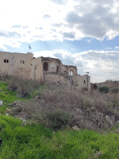 The image captures ancient stone ruins, identified as Antipatris Fortress, on a hillside in Rosh HaAyin, Israel, under a partially cloudy sky. The primary subjects are the dilapidated light-colored stone structures, exhibiting significant damage and structural collapse. The leftmost section of the ruins is a relatively intact wall featuring two small arched window openings and a larger rectangular aperture, with a flagpole displaying a light-colored flag mounted atop this section. To the right, more extensively damaged sections present multiple large arched openings, revealing interior spaces and crumbled masonry. Two black, modern-style lampposts are visible within the ruins, one positioned to the left and another partially obscured on the far right. The foreground consists of a vibrant green grassy slope, which transitions into a dense midground area dominated by dry, brown, woody scrub, thorny bushes, and scattered rocks. The background sky is blue with numerous white and grey cumulus clouds, through which sunlight appears to break in several areas. No individuals are present in the scene.