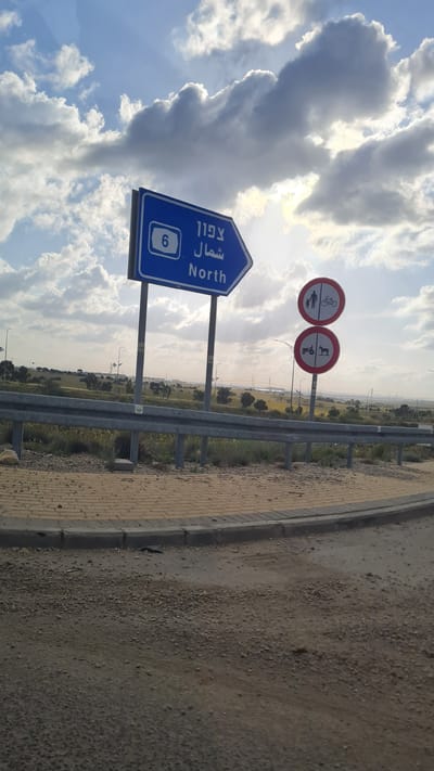 The image depicts a roadside scene in what appears to be the Bnei Shimon Regional Council, Israel. Road signs indicating Route 6 North are visible, along with signs prohibiting pedestrians, cyclists, and tractor-like vehicles. The environment consists of a roadside with a barrier, an adjacent field area, and a sky with scattered clouds. There are no people visible in the image.