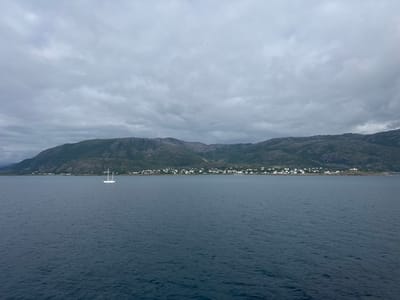 An overcast day looms over a broad expanse of dark, choppy water. In the distance, a rugged, green mountainside slopes down towards a low-lying coastline dotted with a cluster of white buildings, indicating a settlement. A single white sailboat, its mast tall and prominent, floats serenely on the water. The scene is devoid of human presence, creating a sense of quiet solitude. The location is Lødingen, Norway.