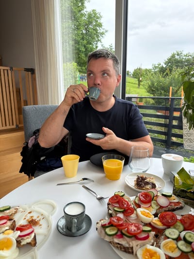 A man is sitting at a white table, drinking coffee from a small cup. He is wearing a dark blue t-shirt. The table is laden with breakfast items including open-faced sandwiches with various toppings like cucumber, tomato, and radish, hard-boiled eggs, a glass of orange juice, and a cappuccino. The scene is indoors, with a large window in the background showing a green, hilly landscape. The man's gaze is directed upwards. There are also some clothes on the chair behind him.
