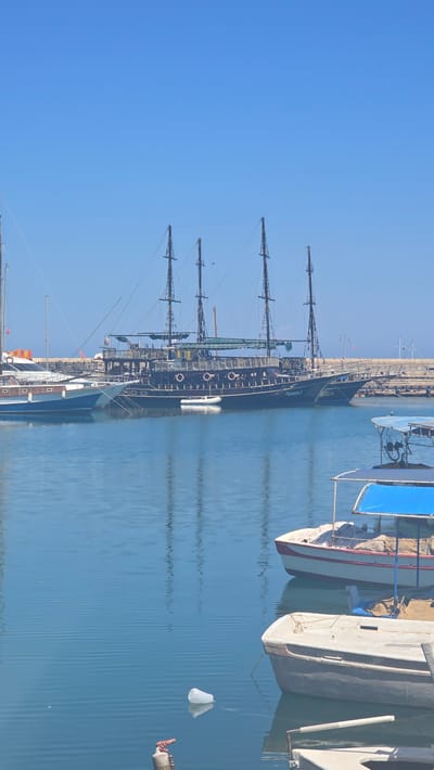 The image shows a harbor scene in Kyrenia, Northern Cyprus. The water is calm and blue, reflecting the clear sky. Several boats are docked in the harbor. The most prominent is a large, dark-colored ship with multiple masts, resembling a pirate ship or a historical sailing vessel. To its left, a smaller white and blue yacht is docked. In the foreground, there are two smaller boats, one with a blue canopy. A white plastic cup is floating in the water. The background shows a breakwater and a clear blue sky.