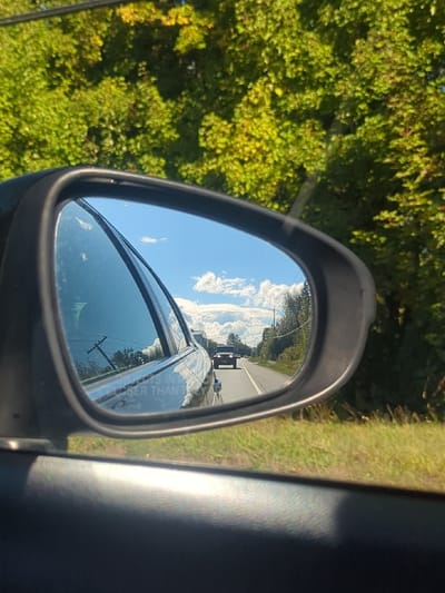 The image presents a view from inside a moving vehicle, centered on its passenger-side rearview mirror. The upper portion of the frame outside the mirror shows dense green and yellowing deciduous tree foliage under bright sunlight. The lower foreground consists of dark, textured interior trim of the car, likely a door panel.

The rearview mirror's reflection displays a two-lane asphalt road extending into the distance under a clear blue sky with scattered white cumulus clouds. A dark, boxy utility vehicle, possibly an SUV or truck, is observed in the far distance, traveling away from the viewer in the same lane with its headlights illuminated. The side of the vehicle from which the photograph is taken, a dark-colored car with a tinted rear window, is partially visible in the mirror's foreground reflection. The warning "OBJECTS IN MIRROR ARE CLOSER THAN THEY APPEAR" is discernible on the mirror's surface. Along the left side of the reflected roadway, two utility poles with wires are present. The immediate roadside within the reflection consists of green grass and some taller vegetation. The scene is consistent with a rural or semi-rural road environment in Maple Ridge, British Columbia, Canada. No people are visible.