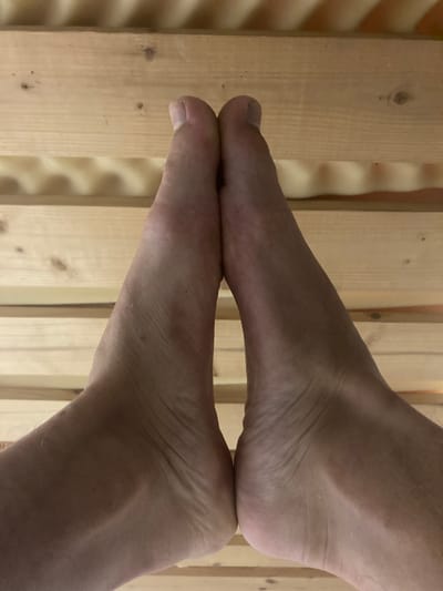 The image displays a close-up of a bare human foot and its reflection, creating the appearance of two feet. The upper foot rests with its toes on the sole of the lower, mirrored foot, forming a symmetrical, almost triangular shape. Both feet have a light skin tone, and details such as toenails, skin texture, and fine hairs are visible.
The feet are positioned against a background of vertical wooden planks, which are light brown or beige with visible grain and knots. The surface on which the feet are resting is highly reflective, creating a clear mirror image of the lower foot. The lighting in the scene appears warm, likely from an indoor artificial source, contributing to the slightly yellowish hue of the wood and skin. There are no discernible shadows that would indicate strong directional natural light. The indoor setting, with its wooden paneling, in Vessigebro, Sweden, could suggest an environment such as a sauna or a room with wooden interior finishes. No text is visible in the image.
