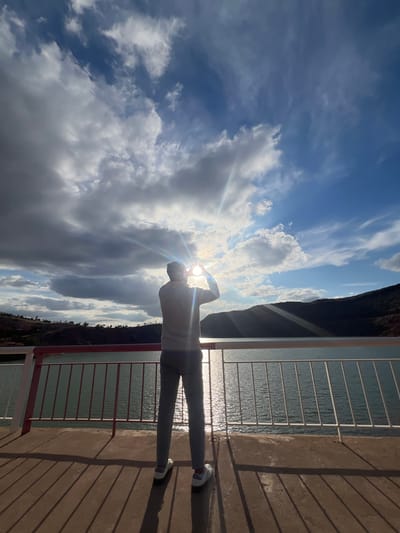 A person stands on an elevated concrete platform in Tabaroucht, Morocco, holding their hands up to frame the sun, which is shining brightly through the clouds. The platform is bordered by a railing with red and white posts. A body of water reflects sunlight. Mountains and vegetation are partially visible in the background. The sky is filled with a mix of dark and light clouds.