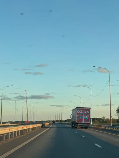 The image shows a road on the outskirts of Bol'shebadrakovo, Russia. An OZON delivery truck is on the road, as well as lamp posts and guard rails. The sky is blue with few clouds.