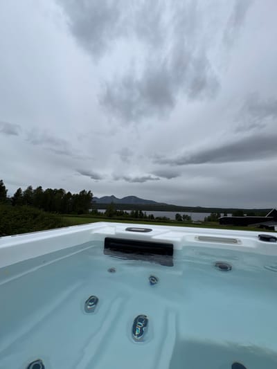The image shows a close-up of the edge of a white hot tub filled with clear water, with several jets visible below the surface. In the background, a serene landscape unfolds under a cloudy sky. A dense line of dark green trees stretches across the middle ground, leading to a calm body of water, likely a lake or fjord. In the distance, the silhouettes of mountains rise against the horizon. The overall scene suggests a tranquil, possibly remote, outdoor setting. The location is identified as Ulvågrenda, Norway.