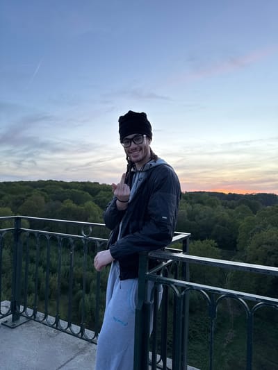 The image captures a young adult standing on what appears to be a high vantage point, possibly a balcony or overlook, above a dense forested valley at sunset. The photograph is oriented vertically, depicting the scene from a tilted perspective.
The subject, likely male, is positioned on the left side of the frame, smiling and looking towards the camera. He wears a black beanie or headwrap, glasses, a black jacket over a grey hoodie, and light grey sweatpants. Dreadlocks are visible peeking from under the beanie. On the left arm of the black jacket, the brand name "RAINS" is clearly visible. On the right leg of the grey sweatpants, there is text written in Arabic script. His right hand is raised, with the thumb and index finger extended in a gesture.
Behind the individual, a panoramic view of a sky painted with sunset hues unfolds. It transitions from soft blues and grays at the top to warm oranges, yellows, and pinks near the horizon, indicating dusk or late evening. Below the colorful sky, a vast expanse of dark green forest covers rolling hills and valleys, stretching into the distance. A dark metal railing with decorative, curved patterns runs across the bottom and right side of the image, separating the viewpoint from the landscape. The ground beneath the railing is a light-colored concrete surface.
The scene conveys a serene and scenic atmosphere, typical of a natural overlook, possibly in Gometz-le-Châtel, France, known for its natural beauty. The person's cheerful expression suggests enjoyment of the view and the moment.