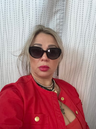 A woman with blonde hair, sunglasses, and bright pink lipstick poses for a photo. She is wearing a red jacket and a low-cut orange top. She has several necklaces layered around her neck, including one with a large cross pendant. The background is a white curtain with a subtle vertical stripe pattern. The image was taken indoors. There is no information provided about the location being in the city of Rome, United States of America.